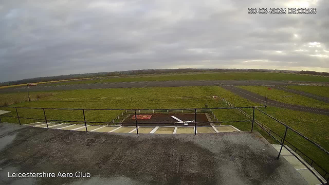 A view from a webcam at the Leicestershire Aero Club. The scene depicts a wide grass field with a runway visible in the background. The sky is overcast with gray clouds. In the foreground, there is a railing and a patch of ground with a white object resembling a model airplane. The landscape is largely open, with hints of vegetation along the periphery.