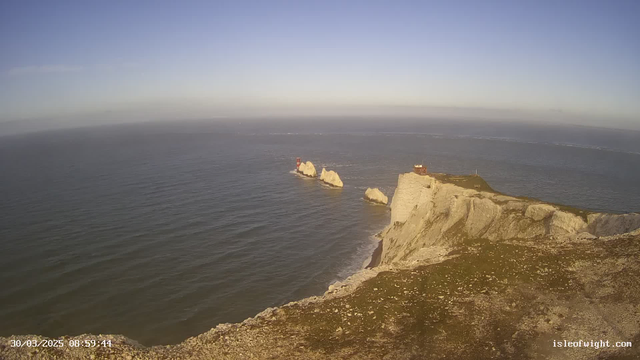 A coastal scene featuring white chalk cliffs that rise steeply from the sea. The water is calm and reflects a clear blue sky with few clouds. In the distance, several small islands are visible, each topped with red and white observation structures. The cliff edge has a grassy area at the top, and the overall atmosphere is serene and picturesque.