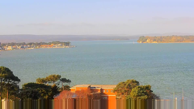 A scenic view of a calm body of water, possibly a bay or estuary, under a clear blue sky. In the foreground, there are several trees and a brown building. On the left side, a marina or shoreline with several boats and buildings can be seen. In the distance, gentle hills can be seen along the horizon.
