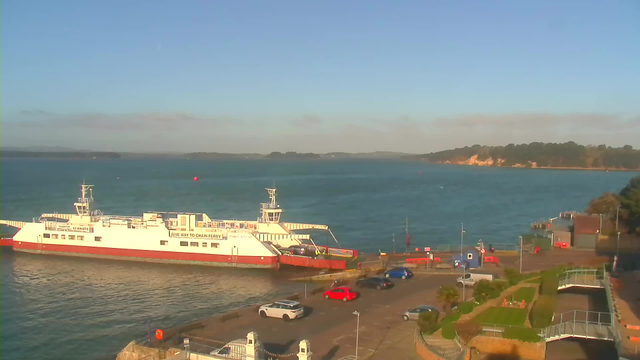 A scenic view of a harbor on a clear day. A white ferry with a red bottom is docked at the pier on the left side of the image. Several vehicles are parked nearby, including red, blue, and grey cars. In the background, calm blue water extends towards the horizon, where a green hillside meets the sky. There are a few buildings and landscaping elements along the waterfront, contributing to the peaceful atmosphere.