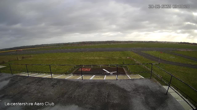 A wide view of an airfield under a cloudy sky. In the foreground, there is a flat rooftop with a railing. Below, there is a section of the airfield featuring a runway and grassy areas. A white-painted aircraft silhouette is on the ground in front of the building. In the distance, there are multiple runways and a sparse landscape with scattered trees. The time displayed at the top right indicates it is 09:00:30 on March 30, 2025.