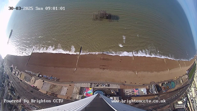 Aerial view of a sandy beach with gentle ocean waves lapping at the shore. The water appears mostly calm with some white frothy surf. Several wooden pier beams extend into the water, and an old, partially submerged pier structure is visible in the distance. The beach is lined with a promenade featuring colorful umbrellas and various structures, including buildings and attractions. The scene is bright with clear blue skies above, indicating a sunny day. The image is timestamped for March 30, 2025, at 09:01:01.