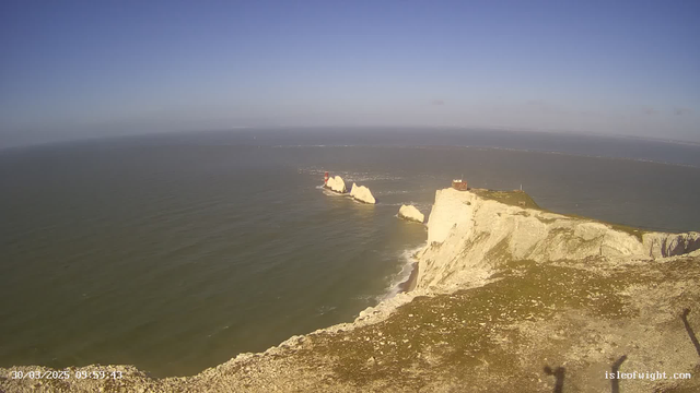 A scenic view of a coastline featuring white cliffs on the right side. In the distance, there are large rock formations protruding from the water, with two lighthouses marked by red and white coloring. The sky is clear and blue, and the ocean is calm with gentle waves. The foreground shows a grassy area with some rocky terrain.