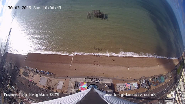 A high-angle view of a beach with golden sand and gentle waves lapping at the shore. There are several people walking along the beach, and a partially submerged pier structure is visible in the water. The shoreline features various colorful beach huts and a promenade lined with buildings. The sky is partly cloudy, and the image timestamp indicates it is a Sunday in March 2025.