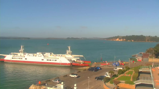 A ferry is docked at a harbor, with a blue sea in the background and a clear sky above. The ferry is white with red accents and is labeled with "Give Way to Chain Ferry." There are several parked cars along the waterfront, with more vehicles nearby. On the right side, there are some green areas and paths leading to a building. The coastline features trees and a sandy area in the distance.