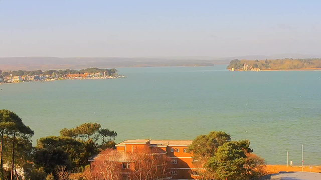 A panoramic view of a body of water surrounded by greenery. In the foreground, there are several trees and an orange building. The water is calm and reflects shades of blue and green. In the background, a small marina with boats is visible along with distant hills. On the right side, a coastal area with a structure is seen on a small island. The sky is clear with pale sunlight illuminating the scene.