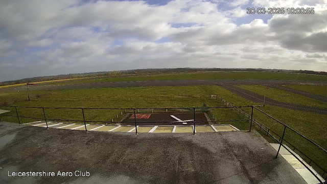 A wide view of an airfield taken from a rooftop vantage point. The scene shows expansive green fields with some patches of grass and a gray tarmac runway. In the foreground, there is a railing along the edge of a flat rooftop, and the ground features a circular area marked by a white object, which may indicate a helipad. The sky above is partly cloudy, with patches of blue peeking through the clouds. The date and time are displayed in the upper right corner.