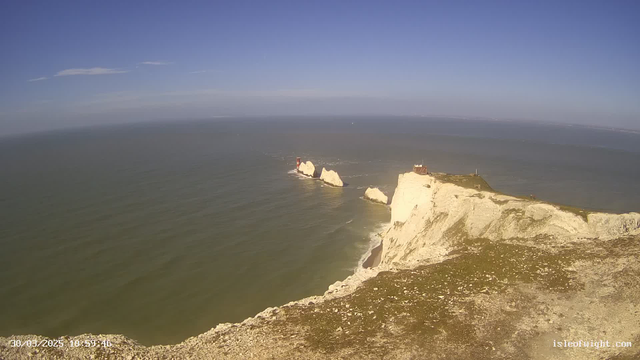 A panoramic view of a coastline featuring white cliffs dropping into the blue sea. In the background, a clear sky with a few wispy clouds can be seen. There are two prominent red lighthouses near the cliff edge, surrounded by rocky formations. The water appears calm with gentle waves. The foreground shows a grassy area on top of the cliffs, while the beach is visible below, leading into the water. The date and time are displayed in the lower left corner of the image.