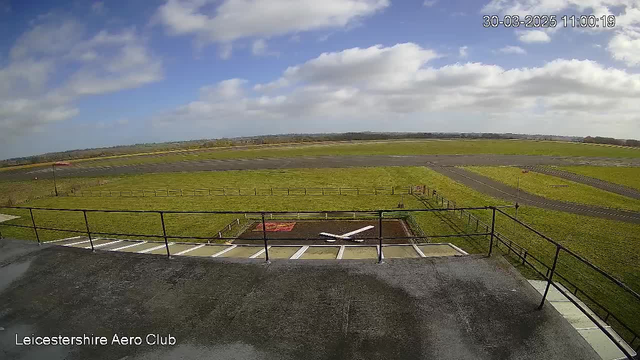 A view from a high vantage point at an aeroclub, showing a large grassy field with scattered patches of yellow and brown. A paved runway extends across the scene, with several clouds in a bright blue sky. In the foreground, there is a railing and part of the building, with a small white airplane silhouette visible on the ground below. Further in the distance, the landscape includes rolling hills and patches of trees along the horizon.