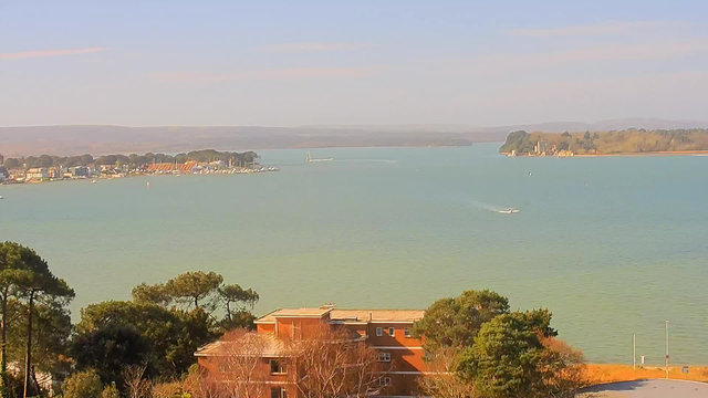 A panoramic view of a calm bay with a light blue-green water surface. In the foreground, there are several residential buildings with a reddish-brown hue surrounded by trees and shrubs. To the left, a marina can be seen, dotted with boats moored along the coast. Gently rolling hills are visible in the background, under a clear sky with soft, scattered clouds. A small sailboat is moving across the water in the center of the bay.