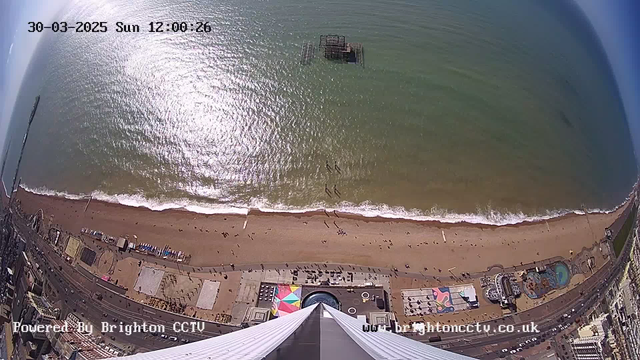 Aerial view of a sandy beach alongside the ocean. Gentle waves lap at the shore, and several people can be seen walking along the beach. In the distance, an old pier structure is partially submerged in the water. Below, colorful beach huts and amusement park attractions are visible, with people scattered throughout the area. The sky is clear, and the sunlight glimmers on the water's surface.