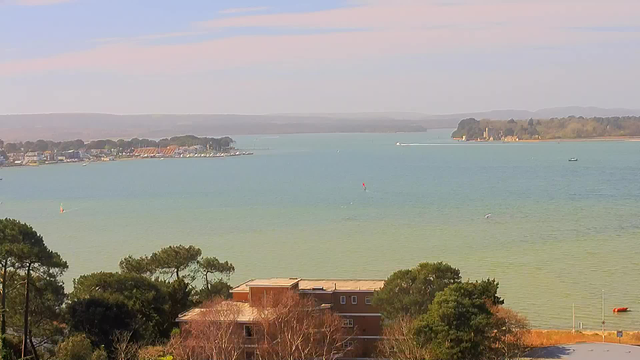 A scenic view of a calm body of water with gentle waves, reflecting shades of blue and green. In the foreground, there are patches of trees, and a building with a flat roof partially visible. To the left, a cluster of houses and a dock can be seen along the shoreline. In the distance, there are boats on the water, and a backdrop of hills under a clear sky with soft clouds.