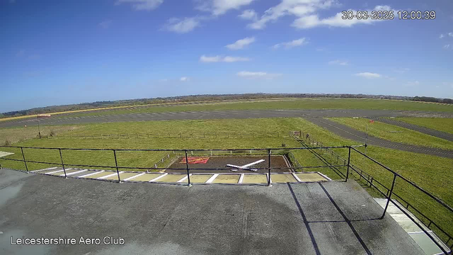 A clear blue sky with a few scattered clouds above a wide open field. In the foreground, there is a flat rooftop with a railing. Below, a green grass field stretches out with a runway in the distance. A marked landing area, featuring a red square and a large white "X," is visible in the center of the field. Several wooden fences outline the area. The scene conveys a tranquil, sunny day at an aerodrome.
