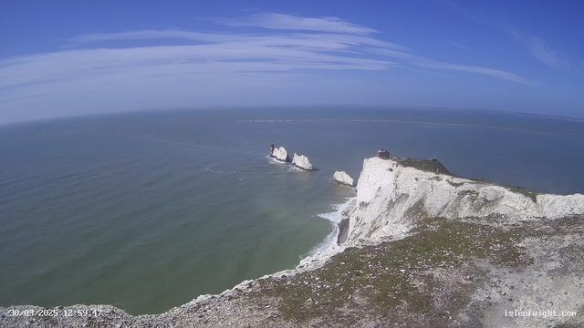 A coastal landscape featuring white chalk cliffs descending to the sea. The sky is bright blue with some wispy clouds. In the distance, there are several rocky formations in the water, with a red lighthouse on one of them. The calm sea reflects the sky and has gentle waves, while some greenery and scattered rocks are visible along the cliff edge.