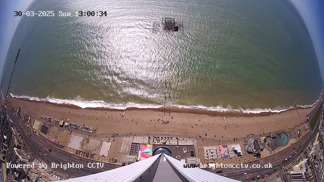 Aerial view of a beach and ocean. The image shows a sandy beach with scattered people enjoying the sun. Gentle waves are lapping at the shore, creating a foamy border. In the foreground, there is a colorful beach setup with geometric patterns. A pier juts out into the sea at the water's edge and further out, a structure is visible in the water. The sky is clear to partially cloudy.