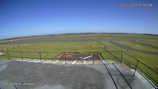 A clear blue sky is shown above a grassy airfield. In the foreground, there is a flat rooftop railing with a view of the runway and taxiways. The ground features a marked helipad with a large white "X" on a brown patch, bordered by a white fence. In the background, a distant line of trees can be seen, indicating the boundary of the airfield.