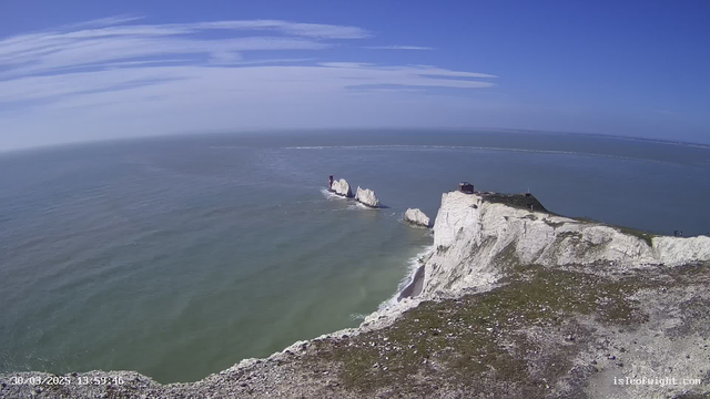 A scenic view of the coastline featuring white chalk cliffs that rise sharply from the sea. The ocean is a mix of blue and green hues, with gentle waves lapping against the shore. In the distance, several rocky formations protrude from the water, with a red lighthouse structure visible on one of them. Above, the sky is mostly clear with a few wispy clouds. The foreground shows grassy land with scattered rocks leading to the cliffs.