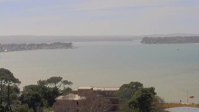 A tranquil coastal scene featuring a calm body of water in varying shades of blue and green. The horizon is lined with gentle hills and a few distant boats are visible on the water. In the foreground, there are trees and a small building near the water's edge. The sky is clear with a few soft clouds, creating a serene atmosphere.