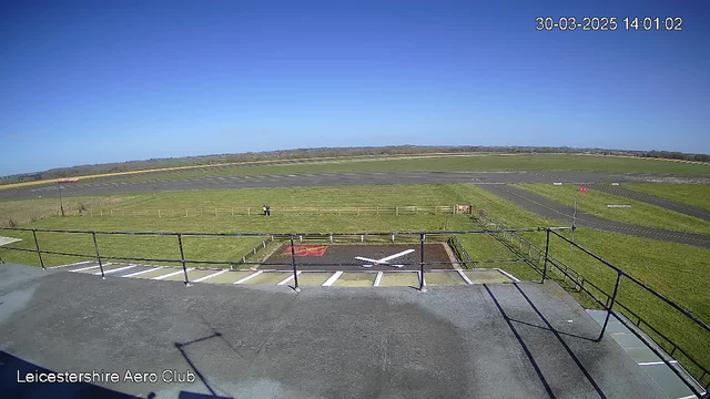 A clear blue sky is visible above a green field, where two people can be seen walking in the distance. In the foreground, there is a flat rooftop area with a railing and markings on the ground that appear to indicate a helipad. There are no aircraft visible in the area. The landscape features a runway in the background, bordered by thin fencing.
