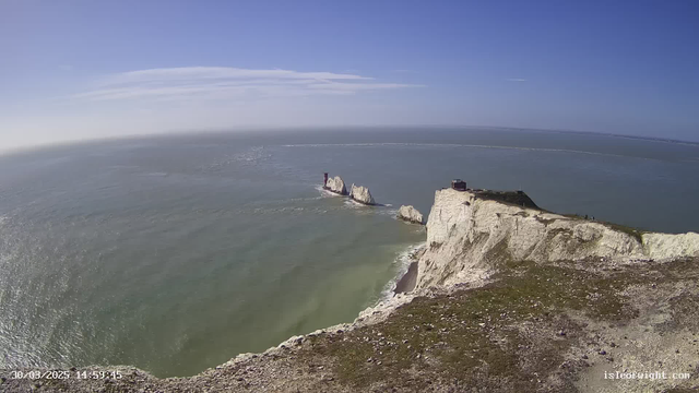 A panoramic view of a coastal landscape featuring white chalk cliffs along the shoreline. The scene includes a calm sea reflecting sunlight, stretching to the horizon under a clear blue sky with a few wispy clouds. In the distance, two prominent rock formations jutting out into the water can be seen, with a red tower-like structure on one of them. The foreground shows grassy areas on top of the cliffs, and there are some small figures visible walking near the edge. The date and time are displayed in the bottom left corner.