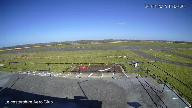 A clear blue sky spans the upper portion of the image, with a few wispy clouds visible. Below, a grassy airfield stretches out, bordered by a wooden fence on the left side. In the foreground, there is a flat surface with a white marking resembling a helicopter landing pad. To the right, two figures are walking along the edge of the grass. Road markings are present on the airstrip, which is visible in the middle distance.