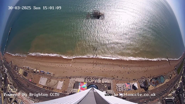 A high-angle view of a beach on a sunny day, with gentle waves lapping at the shore. In the foreground, there is a tall structure, likely a pier or tower, pointing down toward the sandy beach. Scattered groups of people are enjoying the beach. Colorful umbrellas and beach equipment are visible on the sand. In the background, the ocean glimmers under the sunlight, and a partially submerged pier can be seen in the water. A street with buildings runs along the bottom of the image. The time and date are displayed at the top corner.