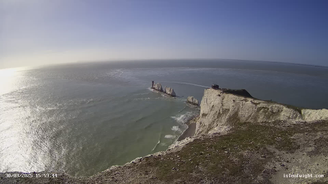 A panoramic view of a coastal landscape shows high white cliffs meeting the ocean. The sea is calm with gentle waves and reflects sunlight, creating a shimmering effect. In the distance, there are a few large rock formations emerging from the water. A small structure is visible on top of the cliff, and the sky above is clear with a few wispy clouds. The scene captures a serene, natural beauty.