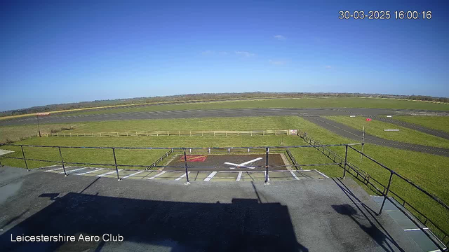 A clear, bright blue sky is visible above a grassy airfield. In the foreground, there is a flat rooftop with a railing. A white "X" marking and a red square are painted on the ground near the edge of the rooftop. Below, the airfield features a long runway with a few painted lines and markings. In the distance, there's a fence lining the airfield perimeter. The overall scene conveys a serene outdoor atmosphere at the Leicestershire Aero Club.