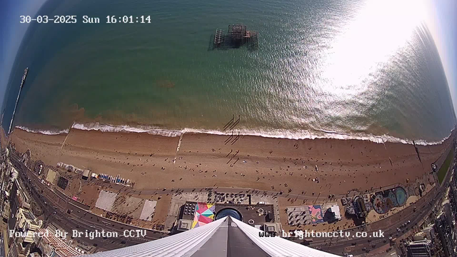 Aerial view of a beach with golden sand and gentle waves lapping at the shore. People are scattered along the beach, engaging in various activities. In the background, the blue-green water extends to the horizon, reflecting sunlight. An old pier is partially submerged in the water. To the right, colorful beach umbrellas and structures can be seen, and a bustling area of buildings and attractions is visible at the bottom of the image. The scene is set on a clear day, with bright sunlight illuminating the landscape. A timestamp in the corner indicates it’s March 30, 2025, at 4:01 PM.