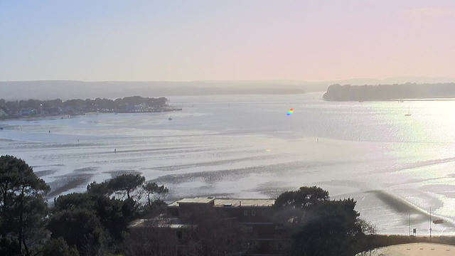 A serene coastal landscape featuring a broad body of water reflecting sunlight. The surface of the water has gentle ripples, creating a shimmering effect. In the distance, there are several small boats scattered across the water, and the shoreline is lined with trees and buildings. The sky transitions from soft blues to a light pink hue near the horizon, indicating early morning or late afternoon light. A slight rainbow shimmer can be seen on the water’s surface, adding a touch of color.