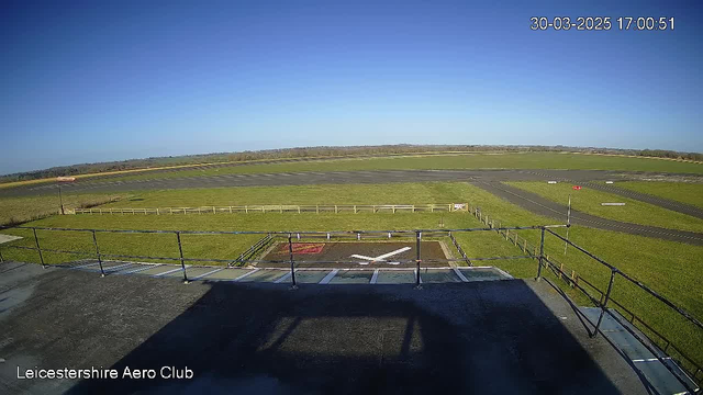 A clear blue sky stretches across the image, with no visible clouds. Below, a grassy airfield is divided into sections, with a marked runway visible. A fenced area is outlined in the foreground, featuring a helipad marked by a white cross. A windsock is positioned to the left, indicating wind direction, while a small red and white signal pole stands at the edge of the runway. The scene captures a tranquil setting, indicating it is a sunny afternoon at the Leicestershire Aero Club.