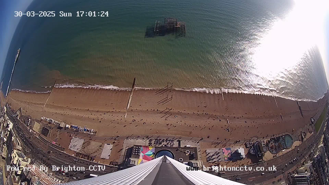 Aerial view of a beach with a sandy shoreline and blue ocean waves. Many people can be seen walking along the beach, with some lying down on the sand. There are structures, including a pier partially submerged in the water and amusement park attractions visible near the beach. The scene is bright, indicating it is daytime, with a clear sky.