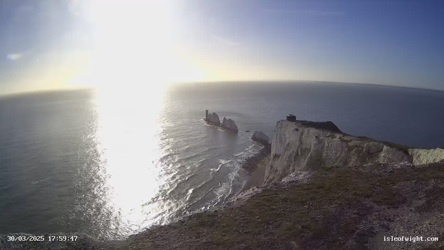 A coastal scene depicting steep white cliffs overlooking the ocean. The sunlight glimmers on the water's surface, creating reflections. In the distance, there are two large rock formations emerging from the sea, with a stack or tower visible on one of them. The sky is clear with a slight gradient from blue to golden near the sun. The foreground includes grass-covered land, leading to the cliff edge.