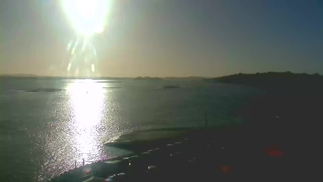 A bright sun shines in a clear sky, casting a warm glow over a calm body of water. The sunlight reflects off the surface of the water, creating a shimmering effect. In the foreground, there are several parked cars along the shoreline, and distant hills can be seen on the horizon. The overall scene conveys a peaceful, serene atmosphere.