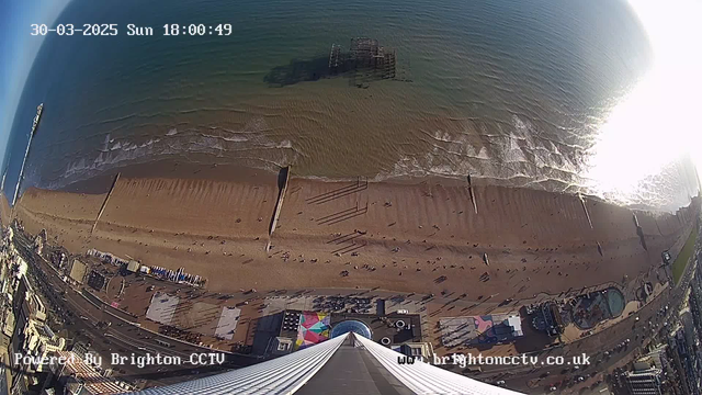 Aerial view of a beach with a sandy shoreline and gentle waves. The ocean appears calm, and there are a few scattered individuals walking along the beach. To the left, a pier extends into the water, with a partially submerged structure visible beneath the surface. Colorful beach huts and parked vehicles are located on the promenade adjacent to the beach. The image shows a clear sky above, indicating a bright day. The date and time are noted in the top left corner.