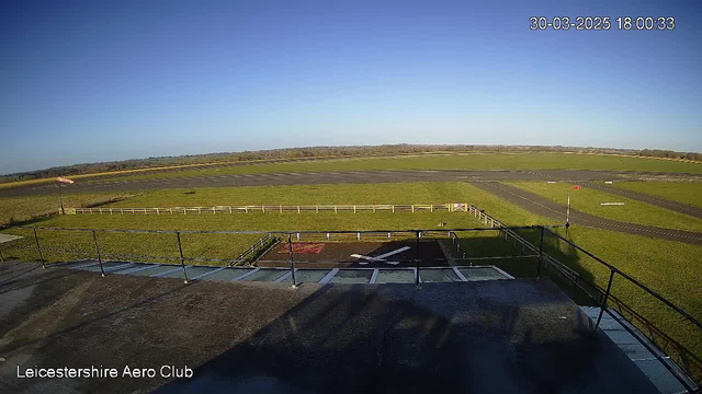 A clear blue sky spans above a wide, open grassy area with a runway. In the foreground, there is a railing with a flat surface just below it. To the left, a small red wind sock indicates the wind direction. The runway is marked with white lines, and there is a small helipad visible on the ground with a white 'H' symbol. The landscape is flat, extending into the distance with trees lining the horizon. The image shows a tranquil outdoor scene at an aero club.