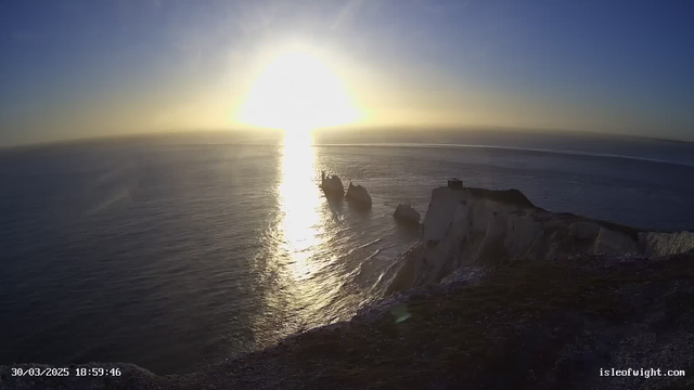 A view of the sea at sunset, with the sun low on the horizon creating a bright golden glow across the water. The shoreline features white cliffs, and several large rocks protrude from the water. The sky transitions from light blue to a deeper blue, indicating the end of the day. A small structure can be seen at the top of the cliff.