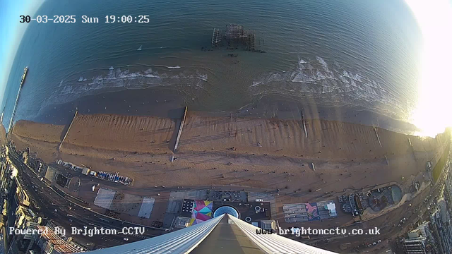 Aerial view of a sandy beach featuring people walking along the shore. Waves gently lap against the sand, creating a shimmering surface. In the foreground, a tall structure is visible, with the shoreline and an old pier extending into the water. Colorful structures can be seen at the bottom of the image, suggesting nearby amusement attractions. The sky is clear with a hint of sunlight reflecting off the ocean's surface.