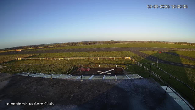 A wide view of an open grass field under a clear blue sky, taken from a high vantage point at the Leicestershire Aero Club. In the foreground, there is a flat rooftop area with a white object resembling a plane's wings. Below, a paved runway runs horizontally across the image, bordered by a white fence on one side. The ground is predominantly green grass, with a few darker patches. There are no visible people or aircraft at this moment. The timestamp indicates 19:00 on March 30, 2025.