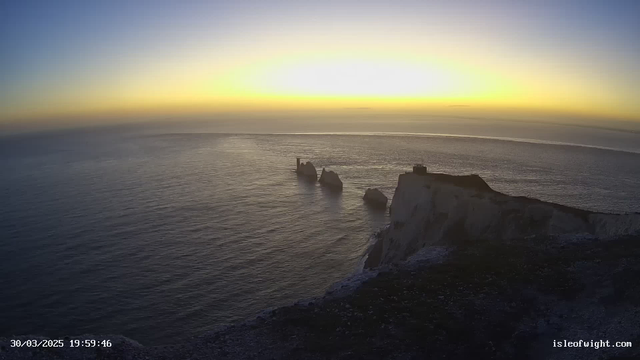 A serene coastal scene at sunset, showcasing a calm sea with gentle waves reflecting hues of orange, yellow, and blue in the sky. Three jagged rock formations emerge from the water, with one featuring a structure on top of it. The cliffs on the right are steep and rugged, leading down to the shoreline. The atmosphere is tranquil and picturesque, evoking a sense of peacefulness.