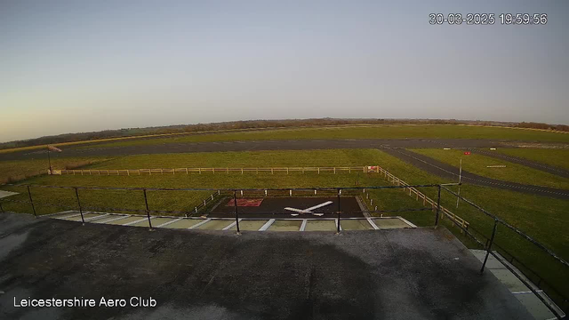 A wide view of an airfield with a green grassy area and a black paved runway in the background. In the foreground, there is a white helicopter landing pad marked with an "X". A wooden fence borders the grassy area, and there is a windsock on a pole indicating wind direction. The sky is clear with a faint gradient of colors suggesting the time is near sunset. The date and time are displayed in the upper right corner of the image.