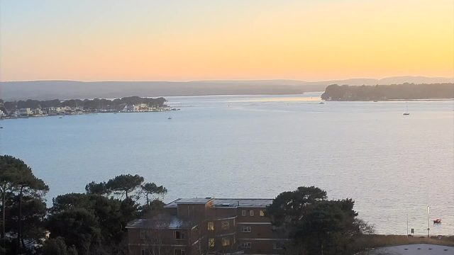 A scenic view of a calm body of water at sunset. In the foreground, there are trees and a low building. The water reflects soft colors, transitioning from light blue to golden hues. Small boats are visible in the water, and a distant shoreline is lined with buildings and greenery. The sky is clear with gentle gradients of color, indicating the end of the day.