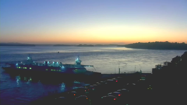 A twilight scene at a harbor. A large ferry boat is docked on the left side of the image, illuminated by blue and yellow lights. The water is calm, reflecting the colors of the sunset, which are shades of orange, pink, and purple. In the foreground, there are several cars lined up, their headlights visible. The background features a shoreline with trees silhouetted against the sky, and distant hills can be seen along the horizon.