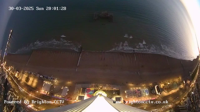 A view from a high angle overlooking a beach and ocean at sunset. The shore is sandy with some structures visible along the beach, including amusement rides and other recreational buildings. The ocean has gentle waves, and a pier extends into the water, partially obscured by the waves. The scene is illuminated by artificial lights along the beach, creating a warm glow in the twilight. The date and time are displayed in the corner, indicating it is March 30, 2025, at 20:01:28.