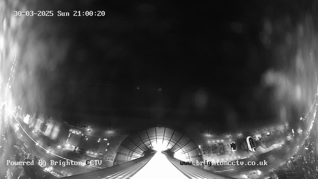 A black and white webcam image showing a dark scene with faint light sources in the background. The foreground features an upward view of a structure with a curved roof, leading towards a bright light at the center, possibly from an opening above. On one side, there are blurred outlines of objects or possibly buildings, with multiple small light points creating an ambient glow. The scene is labeled with the date and time at the top, along with a watermark from Brighton CCTV at the bottom.