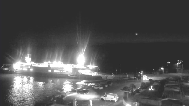 A brightly lit ferry docked at a harbor during the night. The ferry is illuminated with strong lights, reflecting on the water's surface. In the foreground, several vehicles are parked along the waterfront, with some faint lights from additional buildings and structures in the background. The sky appears dark with a hint of starlight.