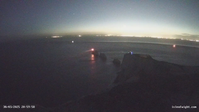 A dark coastal scene at dusk, showing the ocean with a few boats. In the foreground, there are rocky formations emerging from the water. A lighthouse emits a bright light, creating a glowing reflection on the water's surface. The horizon is barely illuminated, with faint lights from distant land visible on the right. The overall atmosphere is calm, with a gradient sky transitioning from deep blue to light near the horizon.