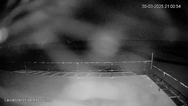 A dimly lit scene captured from a webcam showing a dark outdoor area. There is a railing in the foreground with a few horizontal lines suggesting a fence or barrier. The background features a blurred expanse that appears to be a field or open space, with faint lights in the distance indicating possible structures or roads. The date and time in the corner indicate March 30, 2025, at 21:00:54. Overall, the image has a low visibility due to darkness and possible obstructions in the view.