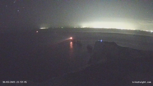 A dimly lit coastal scene at night featuring a body of water. There is a faint glow in the distance suggesting city lights. A small light source, possibly a boat or a buoy, is visible towards the center left. The water reflects some light, creating a subtle shimmer. The shoreline appears dark with indistinct outlines. The date and time are displayed in the lower left corner.