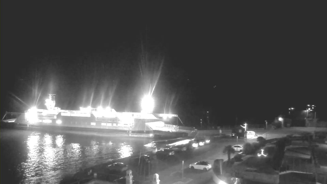 A brightly lit ferry is docked by a quiet waterfront at night. The water reflects the lights, creating glimmers on the surface. In the foreground, several vehicles are parked along the shore, and additional lights can be seen in the background, suggesting a calm and peaceful atmosphere.